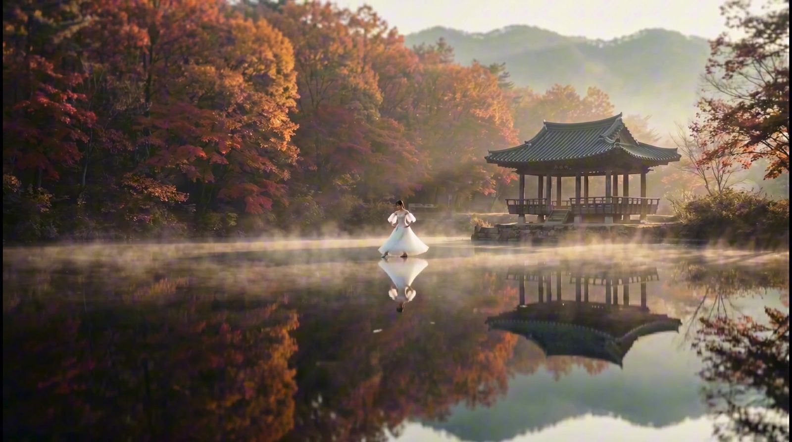 A woman in a white tulle dress stands on a mist-covered lake at dawn, a Korean pavilion behind her among autumn trees.