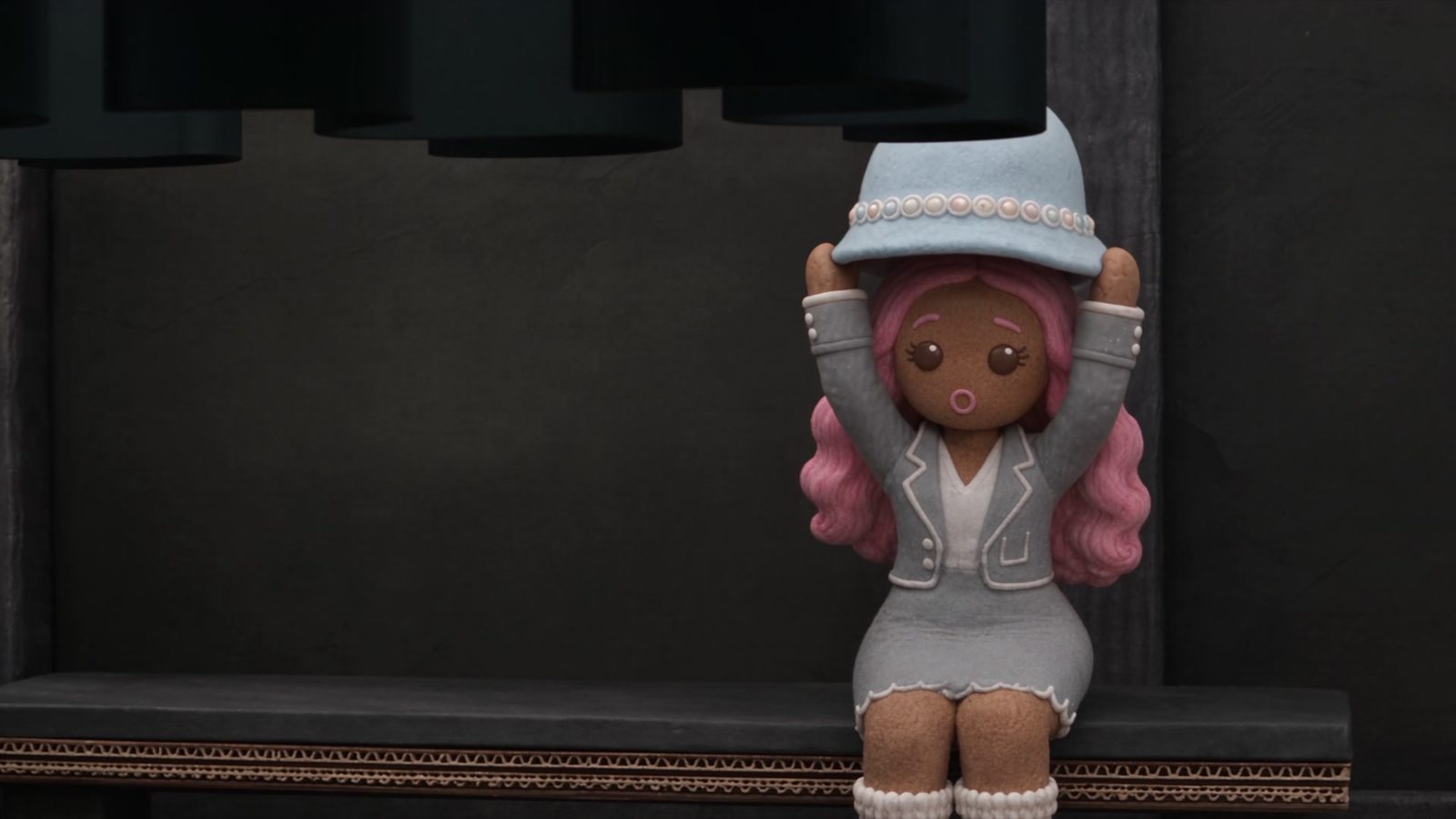 A gingerbread woman with pink hair and a pastel cloche hat sits alone on a steel bench in a dark grey chamber.